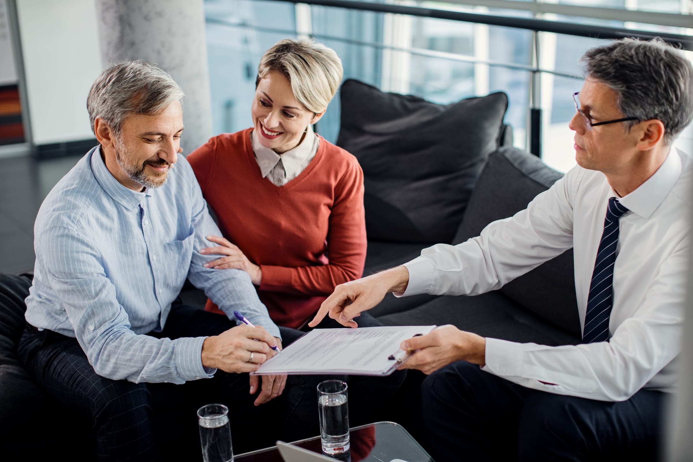 A couple smiling as one of them signs a form held out by an adviser