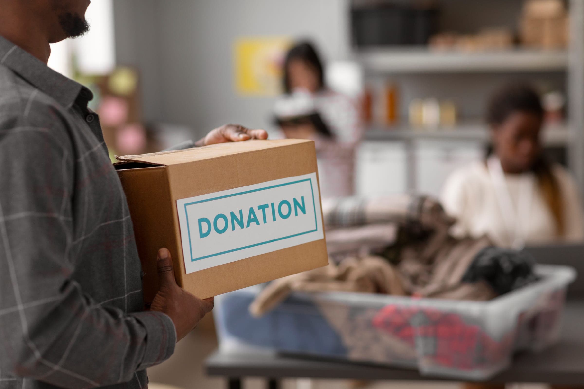 A volunteer carrying a cardboard box labelled Donation, with others sorting clothes behind