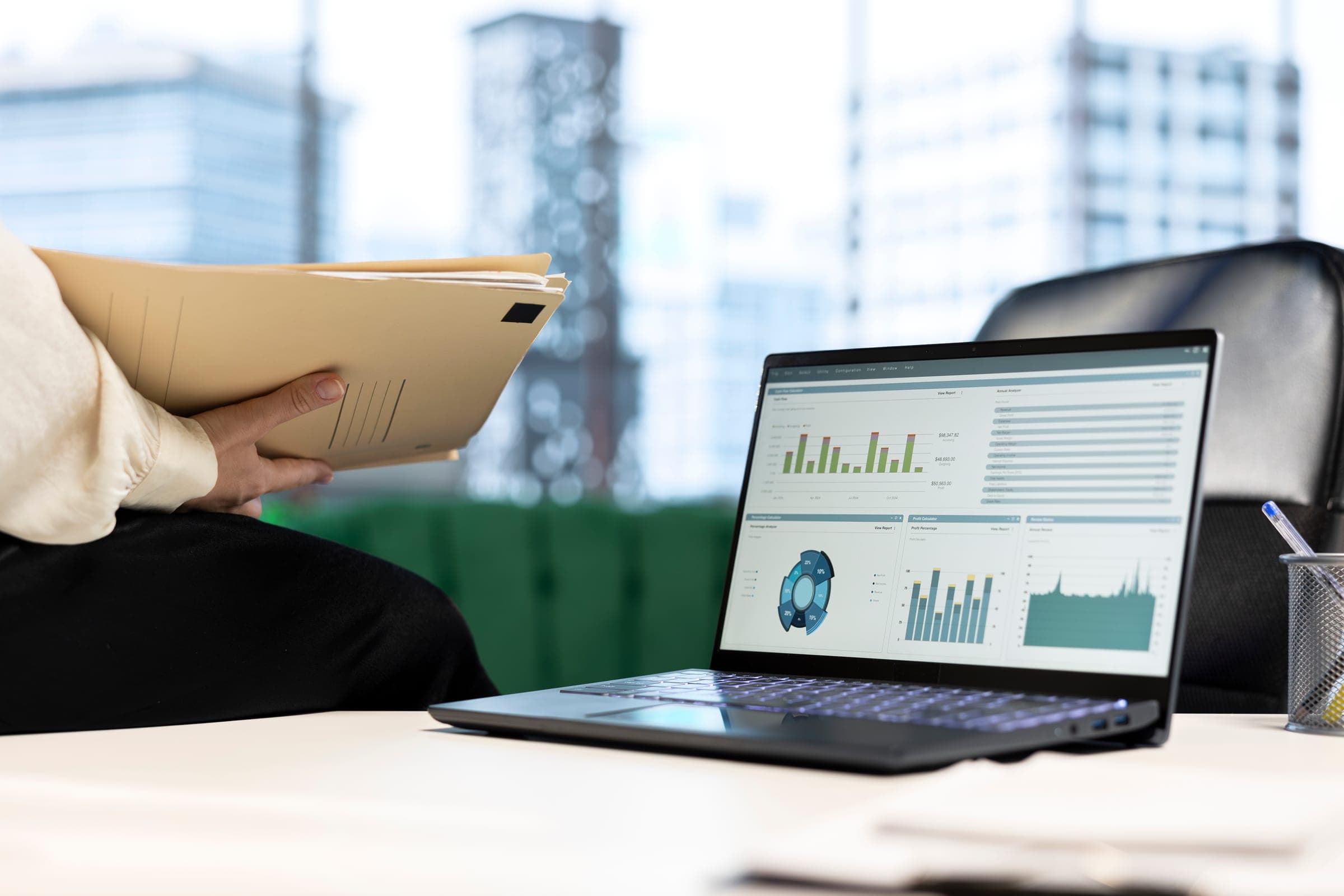 A laptop on a desk showing a dashboard of bar and donut charts, beside a hand holding a folder of papers